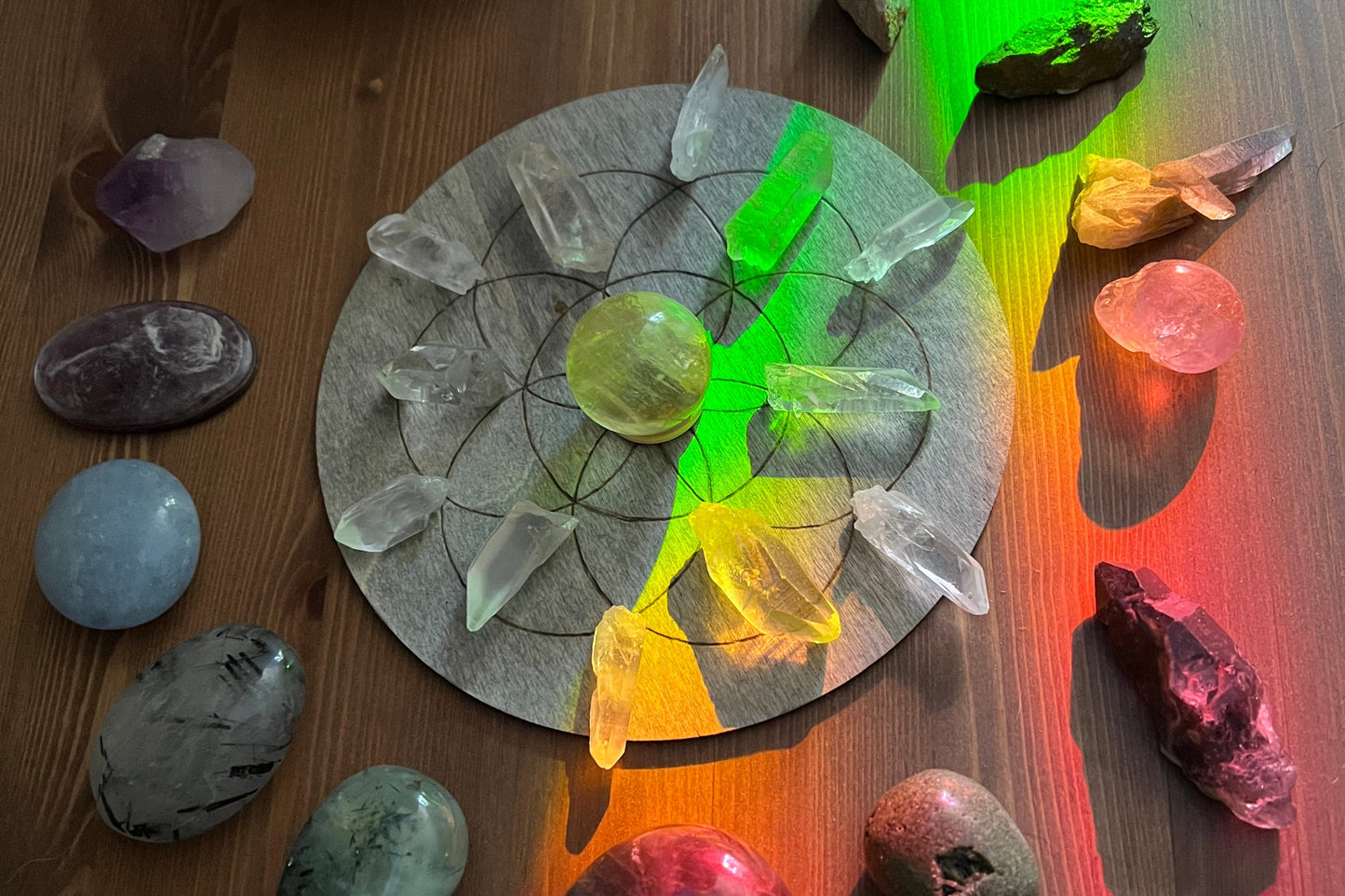A flower of life crystal grid set up with palm stones surrounding it and a rainbow prism light shining over it.