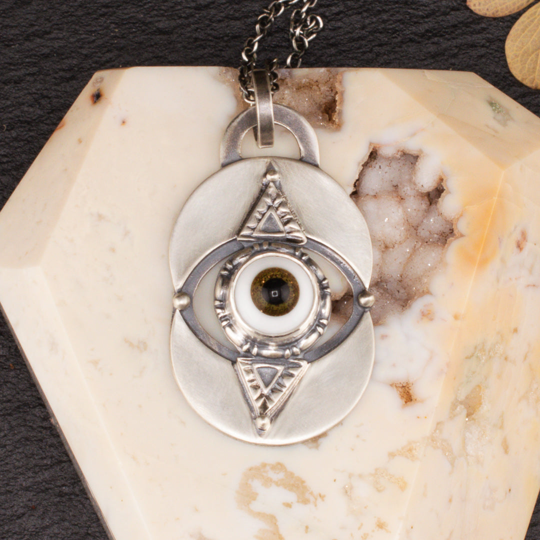 Cloe up of handmade brown glass eye pendant with stamped triangle details set ontop of a Vesica Piscis.