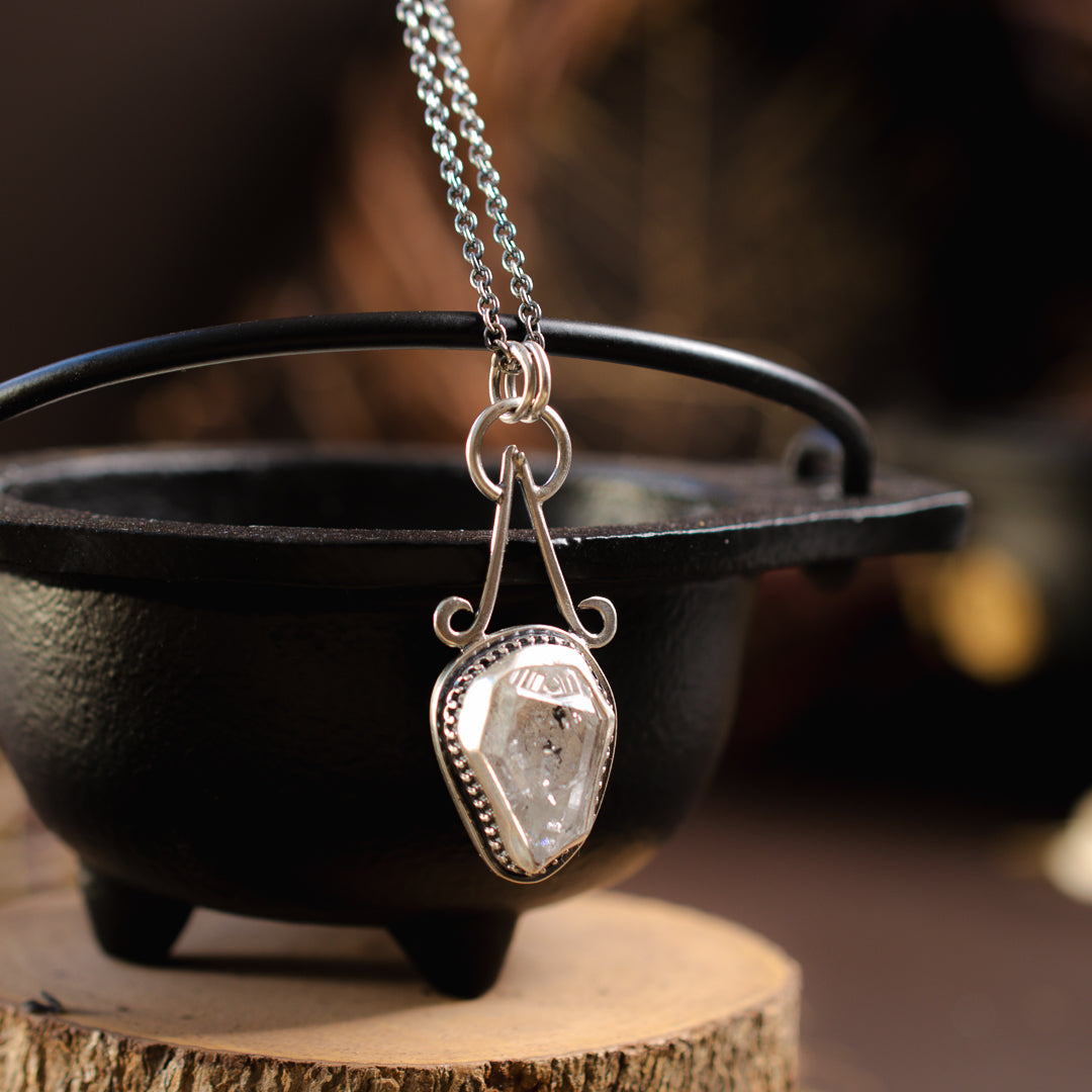 A raw Herkimer diamond set in sterling silver hanging off the side of a cast iron cauldron.