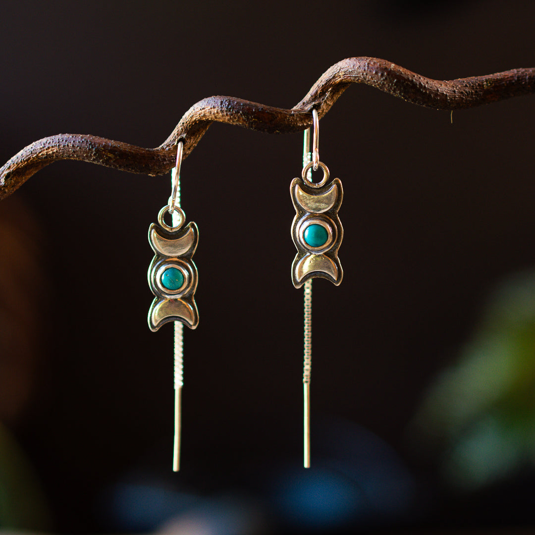 A pair of silver threader style earrings with turquoise accents and a triple goddess symbol, displayed against a backdrop of a twisted metal branch.