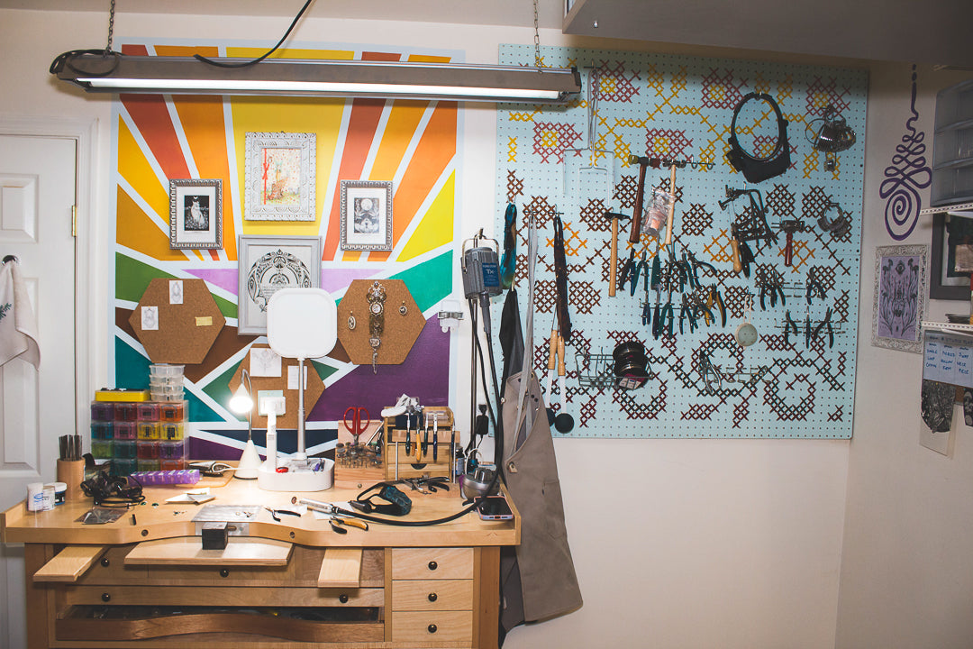 A jeweler's bench in front of a rainbow color mural and a peg board full of jeweler's tools.