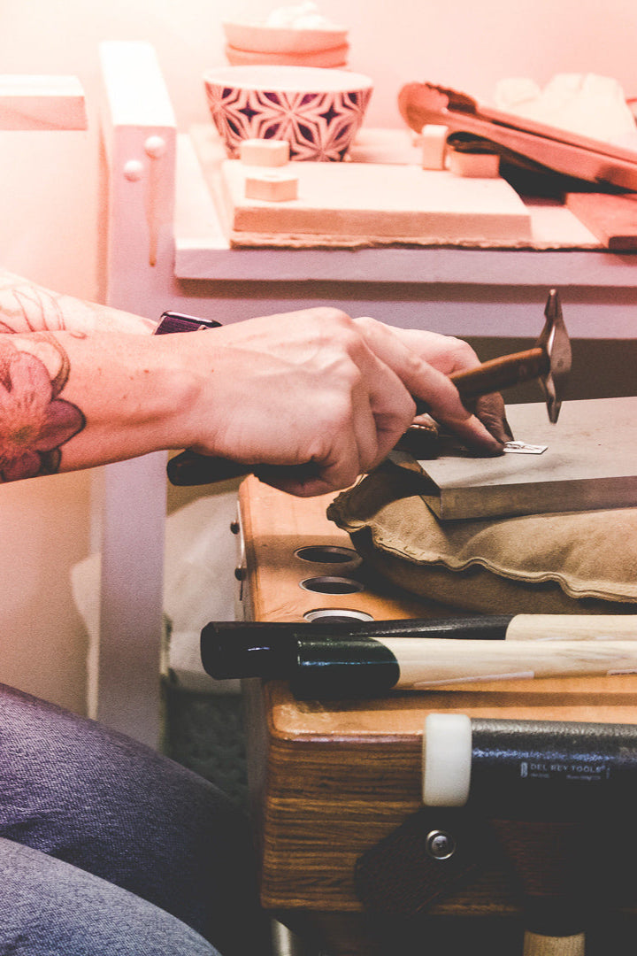 Hands holding a chasing hammer at a wooden anvil getting ready to hammer texture a piece of sterling silver jewelry.