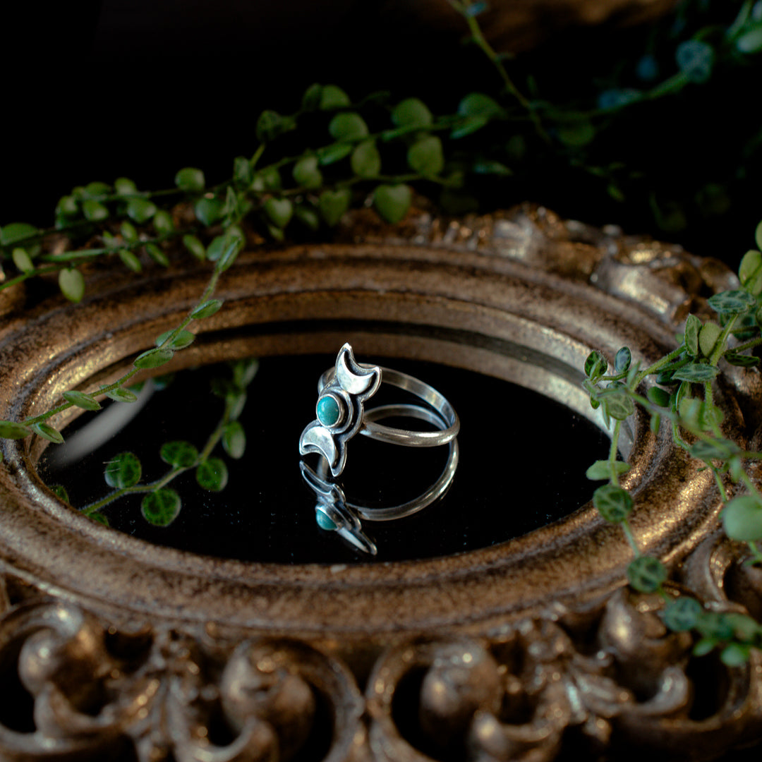 A sterling silver ring with a turquoise stone set in a triple goddess symbol, displayed on a dark reflective surface with greenery in the background.