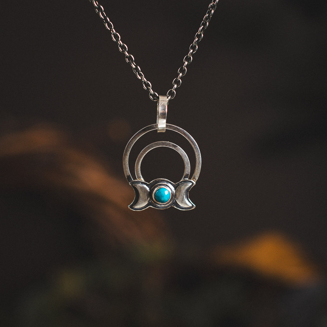 A sterling silver pendant necklace with a triple goddess symbol and turquoise accents, displayed against a dark background with a twisted chain.